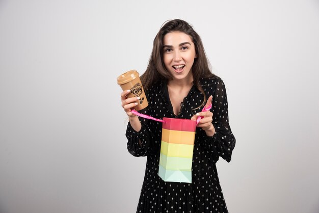 Modelo de mujer joven abriendo una bolsa de la tienda y sosteniendo una taza de café