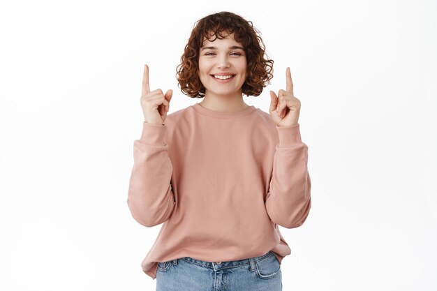 Modelo de mujer joven alegre apuntando con el dedo hacia arriba, sonriendo ampliamente con dientes blancos de pie sobre blanco