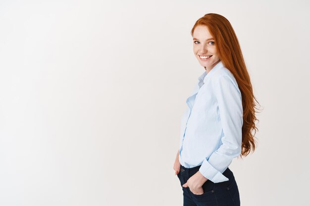 Modelo de mujer joven con cabello pelirrojo y ojos azules gira la cabeza al frente, de pie en camisa de oficina y sonriendo, pared blanca