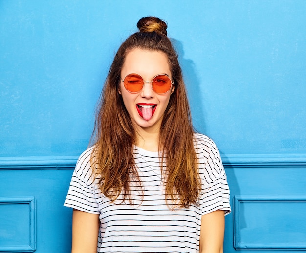 Modelo de mujer joven y elegante en ropa casual de verano con labios rojos, posando junto a la pared azul. Guiñando un ojo y mostrando su lengua