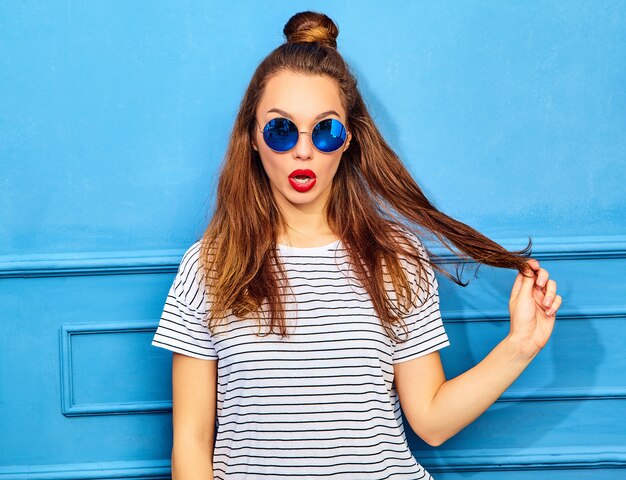 Modelo de mujer joven y elegante en ropa casual de verano con labios rojos, posando junto a la pared azul. Jugando con su cabello