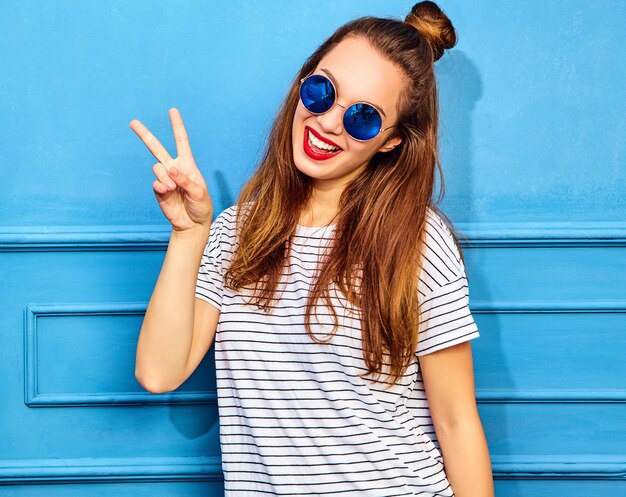 Modelo de mujer joven y elegante en ropa casual de verano con labios rojos, posando junto a la pared azul. Mostrando signo de paz