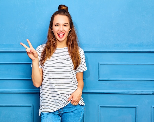Foto gratuita modelo de mujer joven y elegante en ropa casual de verano con labios rojos, posando junto a la pared azul. mostrando su lengua y signo de paz