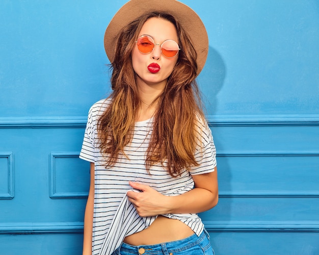 Modelo de mujer joven y elegante en ropa casual de verano y sombrero marrón con labios rojos, posando junto a la pared azul. dando beso de aire