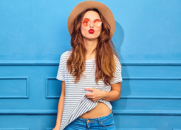 Modelo de mujer joven y elegante en ropa casual de verano y sombrero marrón con labios rojos, posando junto a la pared azul. Dando beso de aire