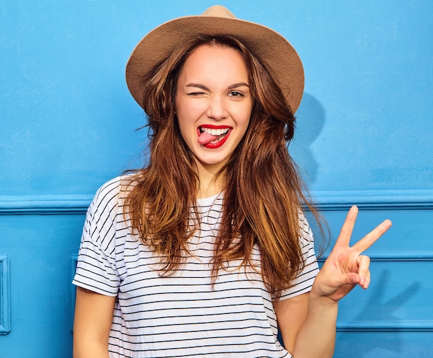 Modelo de mujer joven y elegante en ropa casual de verano y sombrero marrón con labios rojos, posando junto a la pared azul. Guiñando un ojo y mostrando el signo de la paz