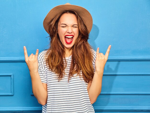Modelo de mujer joven y elegante en ropa casual de verano y sombrero marrón con labios rojos, posando junto a la pared azul. Mostrando el signo de rock and roll