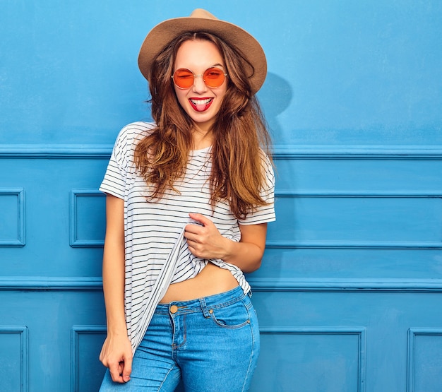 Modelo de mujer joven y elegante en ropa casual de verano y sombrero marrón con labios rojos, posando junto a la pared azul. Mostrando su lengua y guiñando un ojo