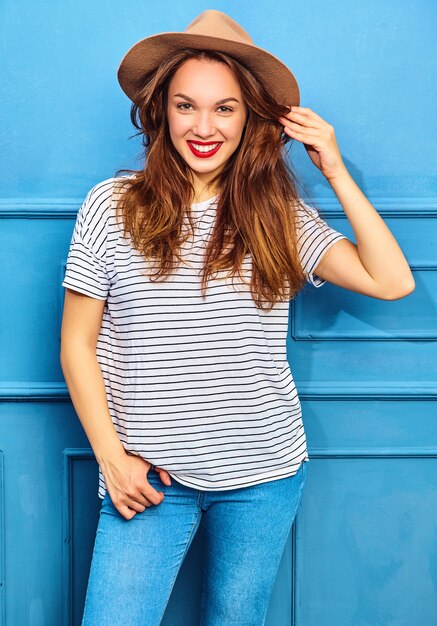 Modelo de mujer joven y elegante en ropa casual de verano y sombrero marrón con labios rojos, posando junto a la pared azul