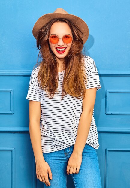 Modelo de mujer joven y elegante en ropa casual de verano y sombrero marrón con labios rojos, posando junto a la pared azul