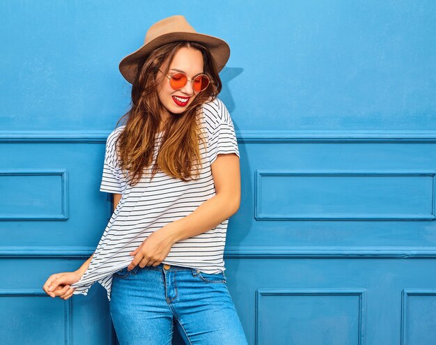 Modelo de mujer joven y elegante en ropa casual de verano y sombrero marrón con labios rojos, posando junto a la pared azul