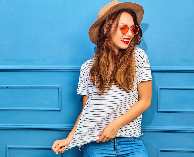 Modelo de mujer joven y elegante en ropa casual de verano y sombrero marrón con labios rojos, posando junto a la pared azul