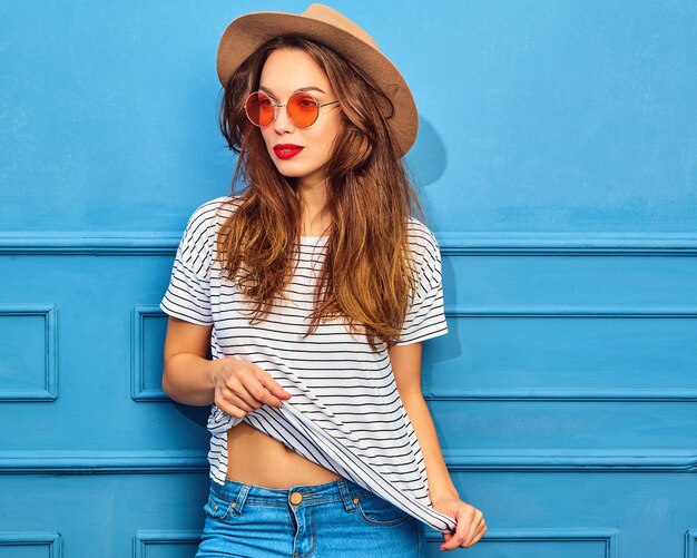 Modelo de mujer joven y elegante en ropa casual de verano y sombrero marrón con labios rojos, posando junto a la pared azul