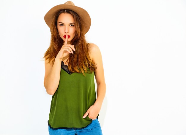 Modelo de mujer joven y elegante en ropa casual de verano verde y sombrero marrón con labios rojos mostrando gesto de silencio con el dedo, aislado