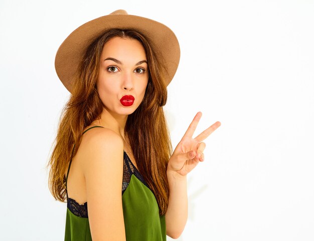 Modelo de mujer joven y elegante en ropa casual de verano verde y sombrero marrón con labios rojos. Mostrando signo de paz y beso