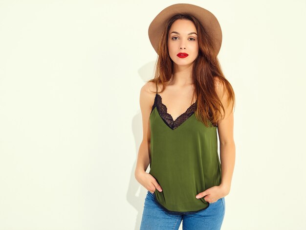 Modelo de mujer joven y elegante en ropa casual de verano verde y sombrero marrón con labios rojos, posando junto a la pared blanca