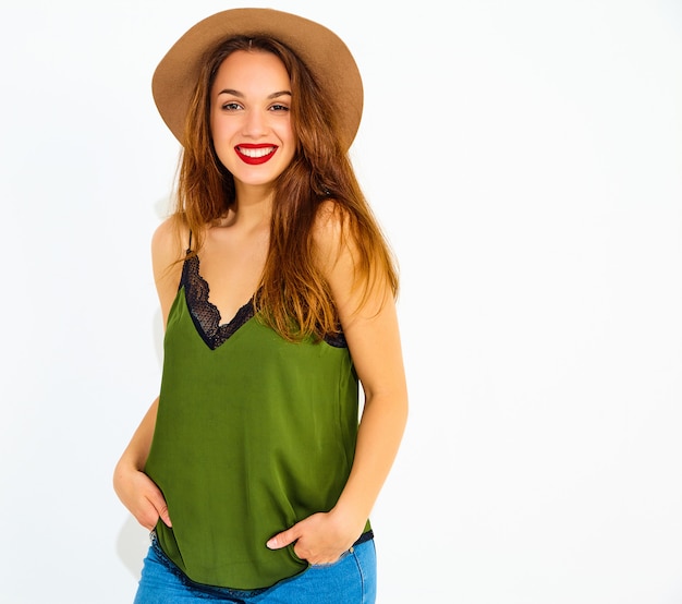 Modelo de mujer joven y elegante en ropa casual de verano verde y sombrero marrón con labios rojos, posando junto a la pared blanca