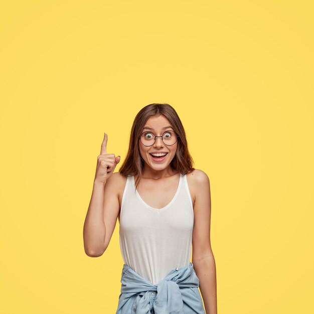 Modelo de mujer joven feliz feliz saliente se siente impresionada por algo hacia arriba, indica con el dedo índice