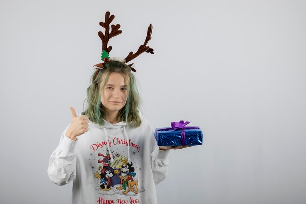 modelo de mujer joven en máscara de cuernos de ciervo sosteniendo un regalo y mostrando un pulgar hacia arriba.