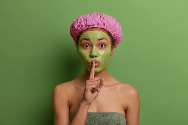 Foto gratuita modelo de mujer joven sorprendida con máscara verde despegable en su rostro, hace un gesto de silencio, dice el secreto de la esteticista, usa gorro de ducha, posa contra una pared vívida concepto de belleza y cuidado facial.