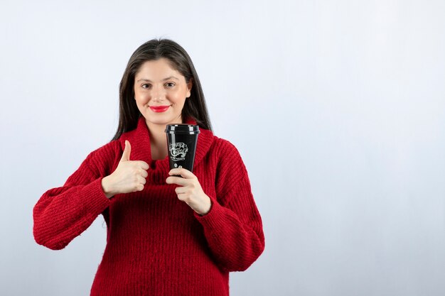 Un modelo de mujer joven en suéter rojo con una taza de café mostrando un pulgar hacia arriba