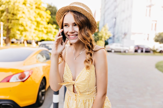 Modelo de mujer ligeramente bronceada con linda sonrisa hablando por teléfono. Chica caucásica entusiasta en vestido amarillo a cuadros posando con smartphone.