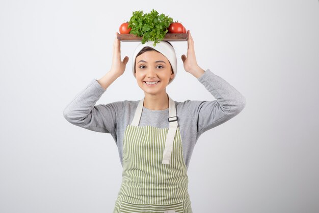 modelo de mujer linda sonriente sosteniendo una tabla de madera con verduras frescas en la cabeza.