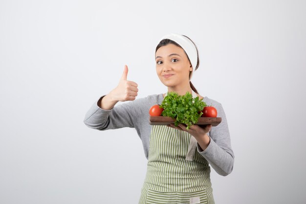 modelo de mujer linda sonriente con una tabla de madera de verduras frescas mostrando un pulgar hacia arriba.