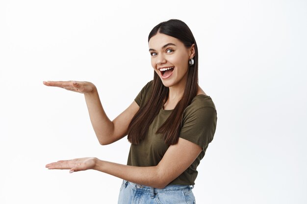 Modelo de mujer morena alegre con espacio de copia vacío para su logotipo o anuncio de producto, haciendo gesto de caja grande, pared blanca