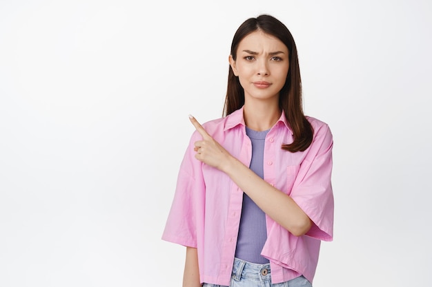 Modelo de mujer morena escéptica apuntando a la esquina superior izquierda frunciendo el ceño y luciendo decepcionada haciendo preguntas con fondo blanco de cara confusa