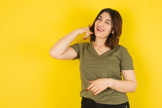 Modelo de mujer morena de pie y mostrando gesto de llamada telefónica contra la pared amarilla