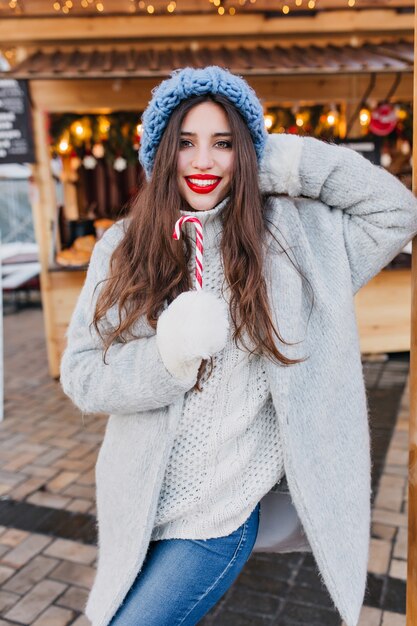 Modelo de mujer riendo con cabello castaño oscuro comiendo dulces navideños en un día frío. Retrato de feliz morena europea en abrigo gris y guantes blancos posando con piruleta en la mañana de invierno.