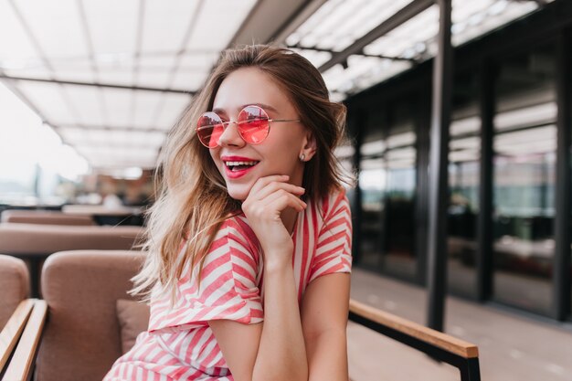 Modelo de mujer romántica en traje de rayas mirando a otro lado con una sonrisa. Foto interior de agraciada dama caucásica relajándose en un café con un interior acogedor.