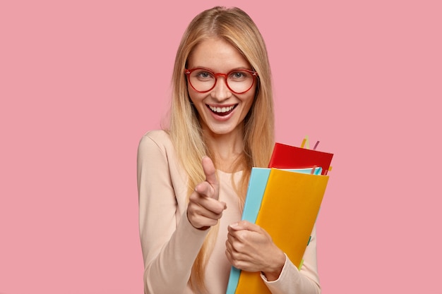 Modelo de mujer rubia alegre lleva gafas redondas, hace gesto de pistola, sostiene libros de texto