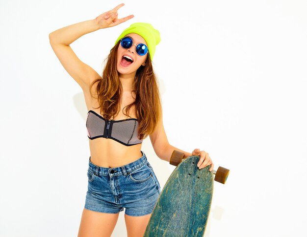 Modelo de mujer sonriente elegante joven en ropa de traje de baño casual de verano y gorro amarillo posando con escritorio de longboard, mostrando el signo de rock and roll. Aislado en blanco