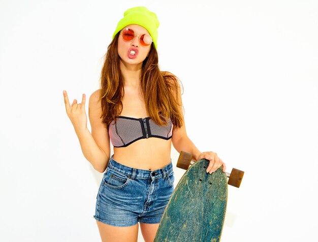 Modelo de mujer sonriente elegante joven en ropa de traje de baño casual de verano y gorro amarillo posando con escritorio de longboard, mostrando el signo de rock and roll. Aislado en blanco