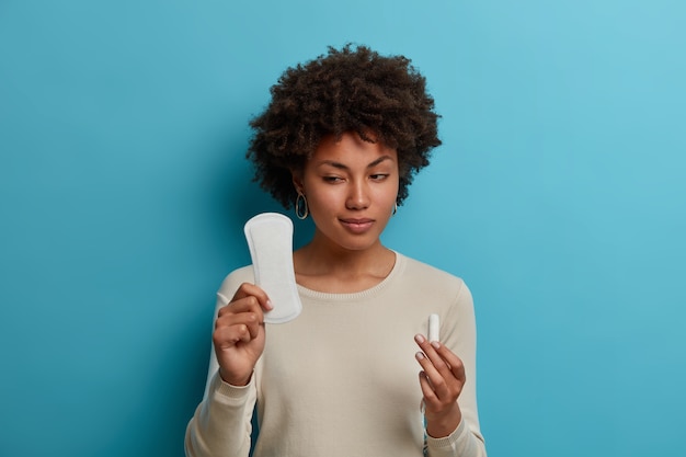 Modelo de mujer vacilante duda al elegir entre compresa y tampón, tiene mirada sospechosa, prefiere algún producto higiénico, vestida con jersey casual. Concepto de menstruación