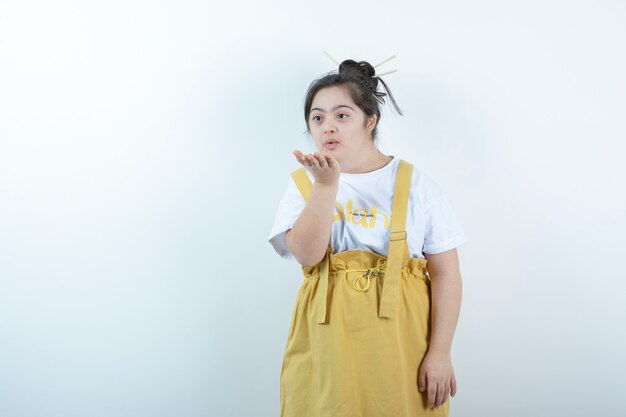 Modelo de niña bonita de pie y soplando un beso al aire contra la pared blanca.