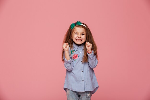 Modelo de niña sonriente en aro de pelo y ropa de moda que expresa felicidad gesticulando con los puños cerrados