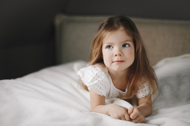 Modelo de niño hermoso está acostado sobre la manta blanca en la cama y mirando hacia un lado