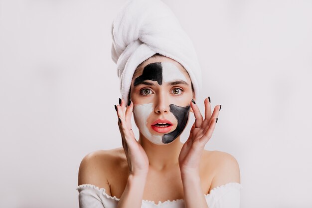 Modelo de ojos verdes después de la ducha hace mascarilla de arcilla blanca y negra. Chica con toalla blanca en la cabeza