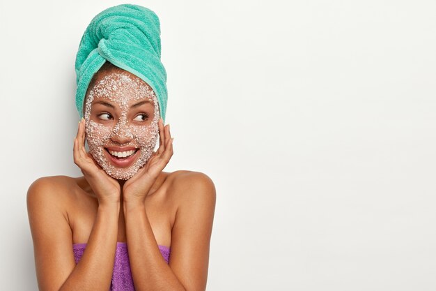 El modelo de piel oscura de aspecto agradable se lleva las manos a la cara, aplica mascarilla exfoliante, gránulos de sal de mar blanca, mira a un lado, está de buen humor