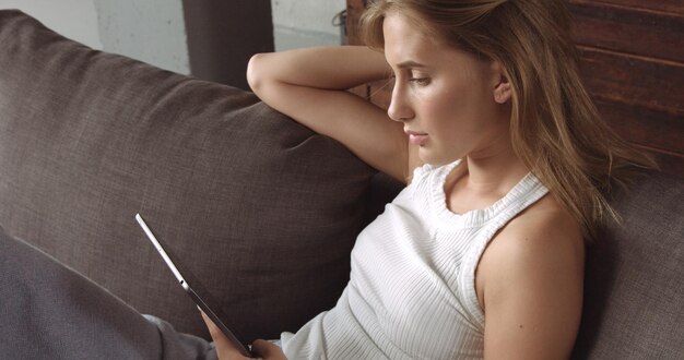 Modelo rubia caliente leyendo en su tableta mientras se sienta en un sofá gris oscuro cubierto con un tiro suave