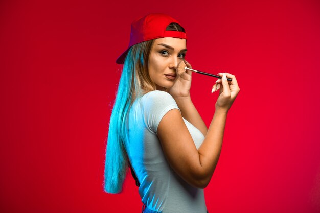 Modelo rubio con gorra roja y aplicando maquillaje.