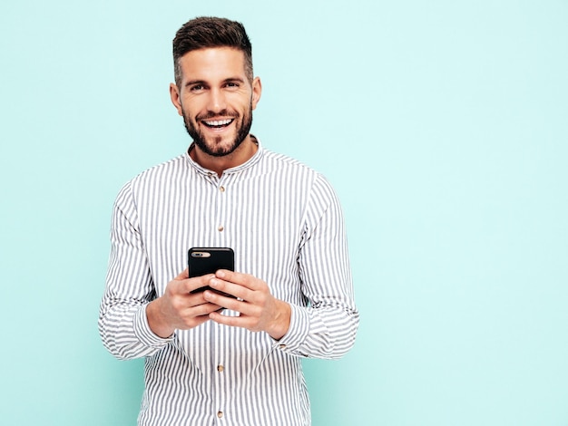 Modelo sonriente guapo Hombre elegante sexy vestido con camisa y jeans Hombre hipster de moda posando junto a la pared azul en el estudio Sosteniendo teléfono inteligente Mirando la pantalla del teléfono celular Usando aplicaciones