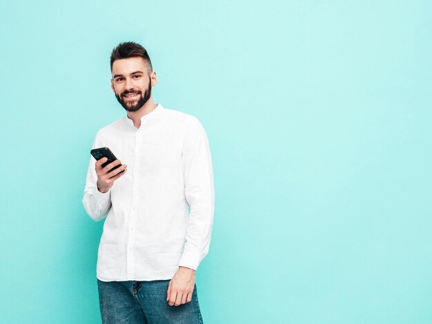 Modelo sonriente guapo Hombre elegante sexy vestido con camisa y jeans Hombre hipster de moda posando junto a la pared azul en el estudio Sosteniendo teléfono inteligente Mirando la pantalla del teléfono celular Usando aplicaciones