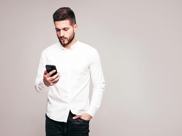 Modelo sonriente guapo Hombre elegante sexy vestido con camisa y jeans Hombre hipster de moda posando sobre fondo gris en el estudio Sosteniendo teléfono inteligente Mirando la pantalla del teléfono celular Usando aplicaciones