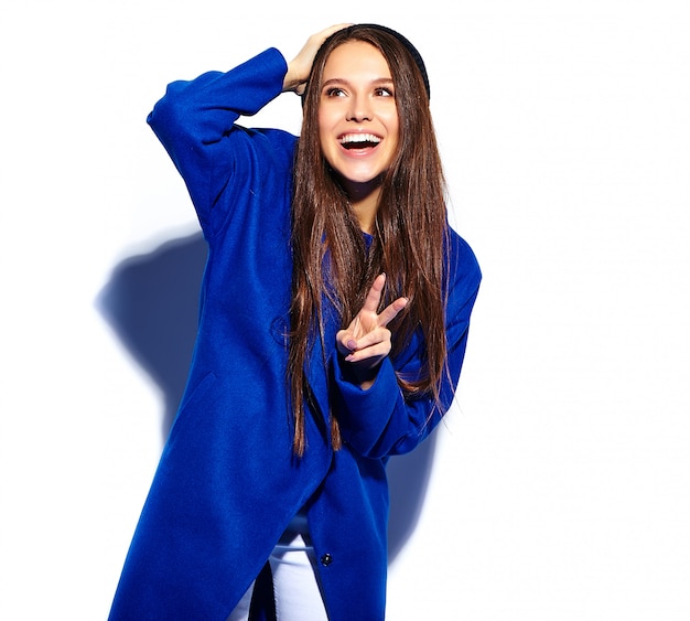 Foto gratuita modelo sonriente hermoso de la mujer morena del inconformista en el abrigo azul elegante aislado en blanco. mostrando signo de paz