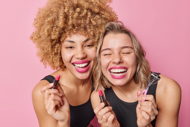 Las modelos femeninas alegres aplican una sonrisa lisptick ampliamente, se divierten mientras se maquillan, se preparan para la fiesta, se ríen felizmente, usan camisetas negras aisladas sobre fondo rosa. Mujeres y procedimientos de belleza.