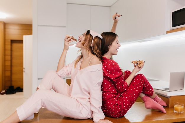 Modelos femeninos atractivos en calcetines lindos sentado en la mesa de madera en la cocina. Chicas refinadas en pijama disfrutando de pizza de queso en casa.